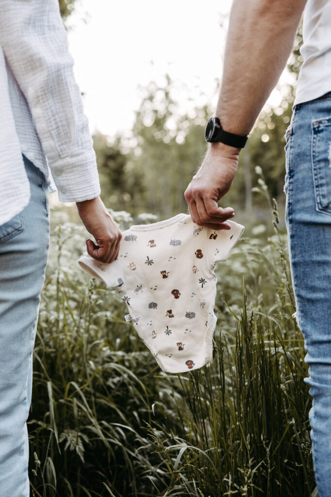 Eltern halten gemeinsam einen Baby-Body mit Tiermotiven in der Wiese