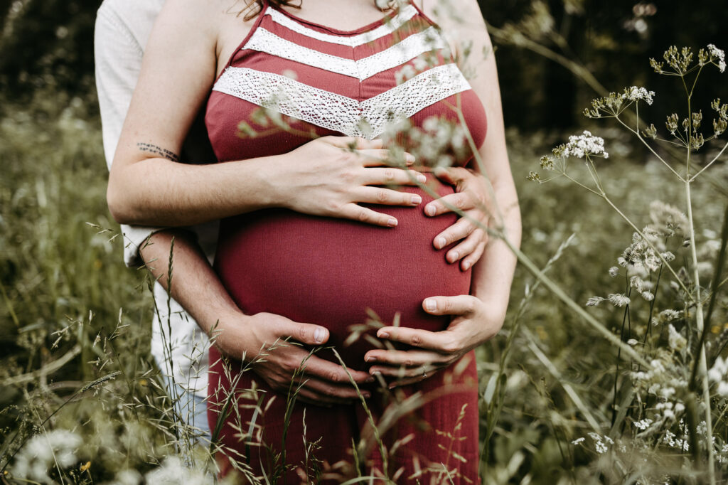 Detail: Hände beider Partner auf dem Babybauch, rotes Kleid, Wildblumenwiese