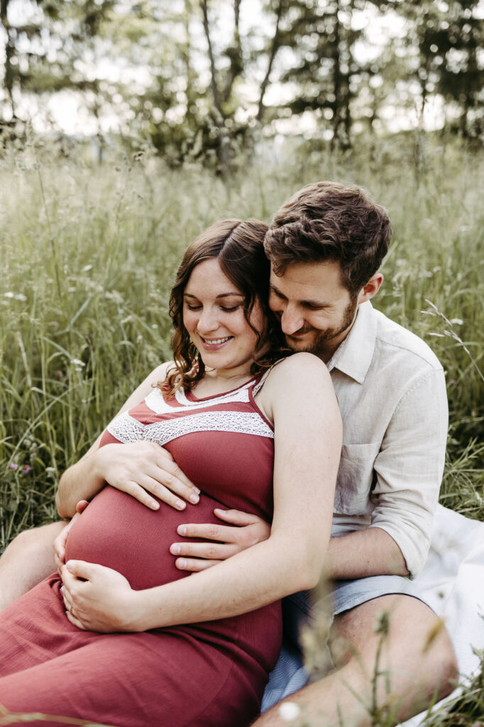 Paar sitzt im hohen Gras, sie in rotem Kleid lächelnd, er hält von hinten den Babybauch, Waldrand im Hintergrund