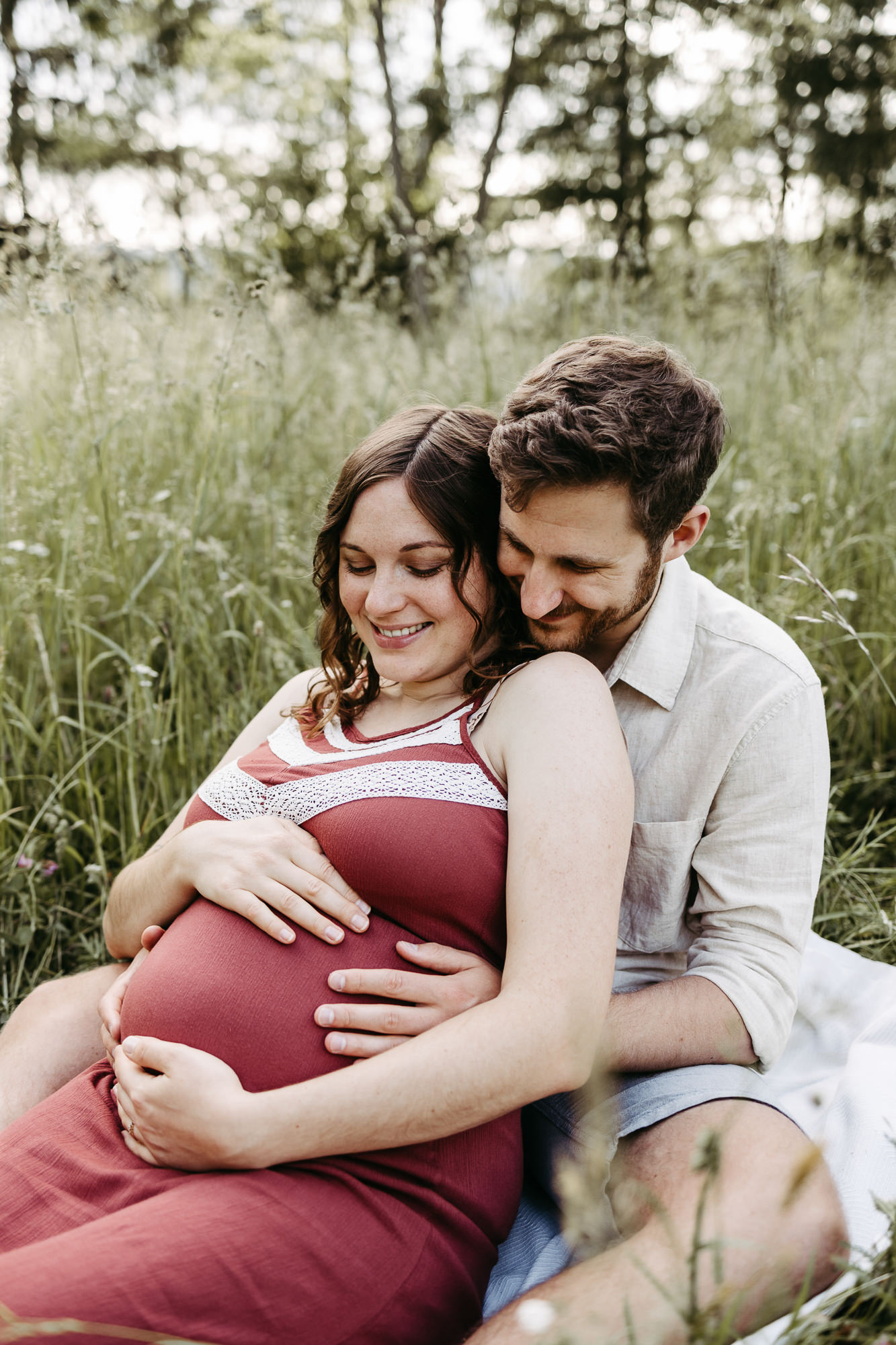 Paar sitzt im hohen Gras, sie in rotem Kleid lächelnd, er hält von hinten den Babybauch, Waldrand im Hintergrund