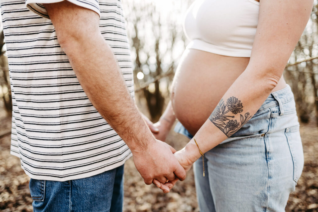Detail: Händehaltendes Paar von der Seite, Babybauch und Blumentattoo im Hintergrund erkennbar