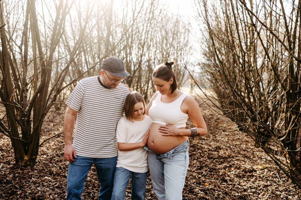 Familie geht über einen herbstlichen Laubweg, Papa und Tochter schauen lächelnd auf den Bauch