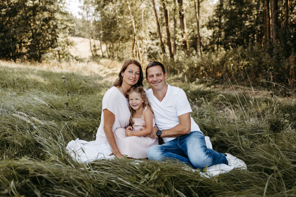 Kleine Familie sitzt lächelnd auf einer Decke in der Wiese, Mädchen im rosa Kleid zwischen den Eltern