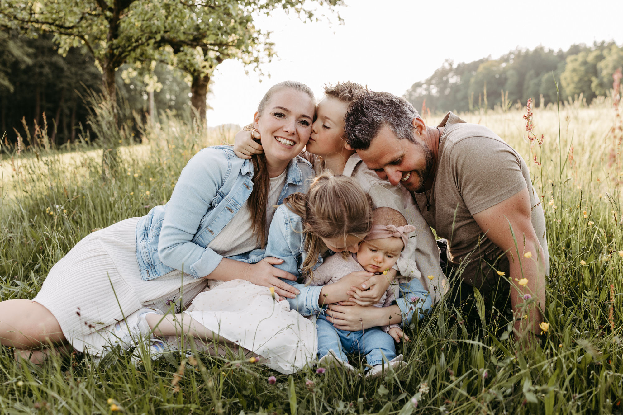 Familie kuschelt sich im Gras zusammen, Sohn küsst die Mutter auf die Wange, Tochter küsst das Baby