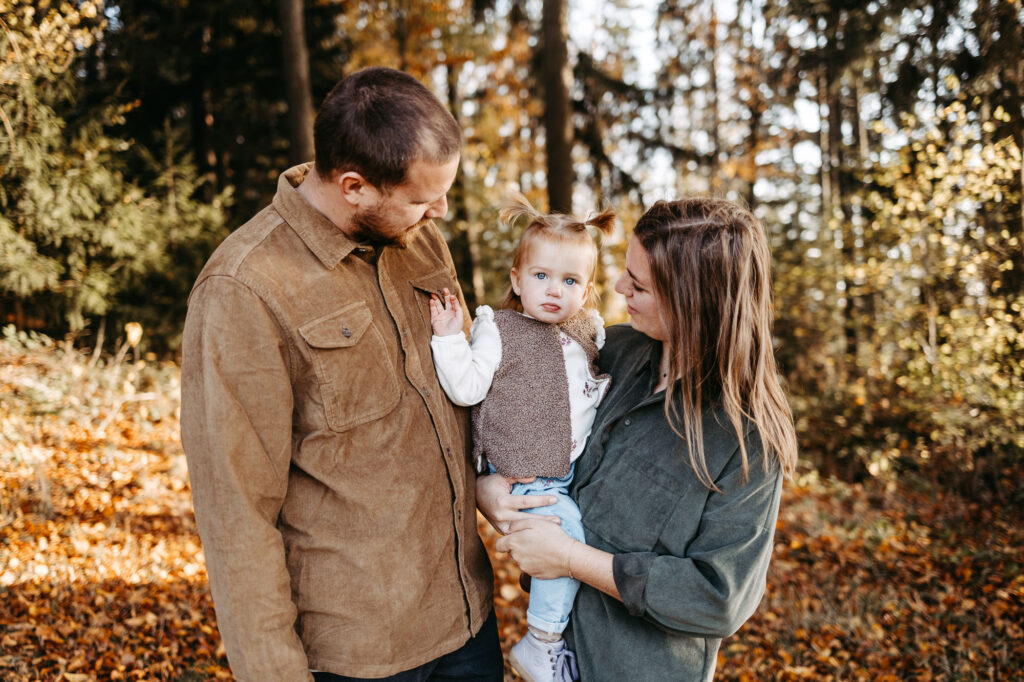 Familienporträt: Eltern blicken lächelnd auf ihre Tochter, goldener Herbstwald mit Laub