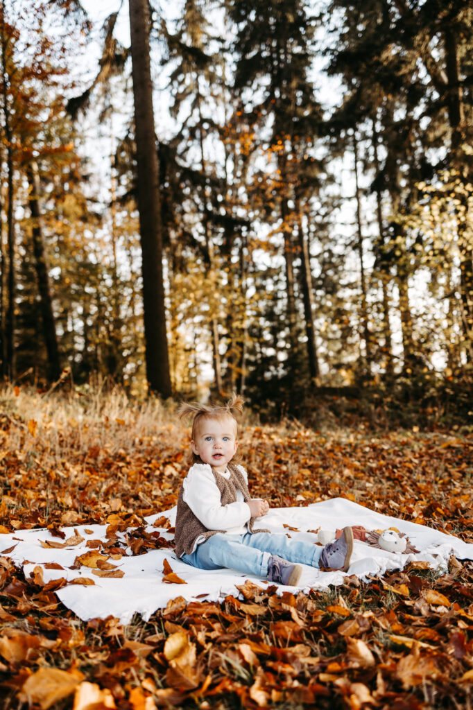 Kleines Mädchen sitzt allein auf einer weißen Picknickdecke, umgeben von Herbstlaub