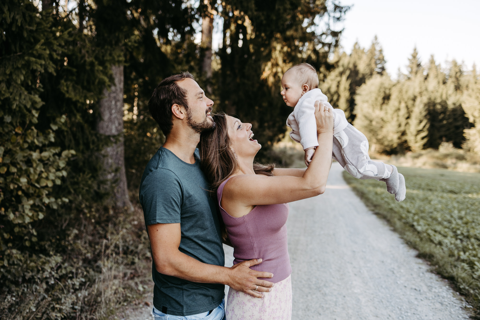 Mutter hebt lachend ihr Baby in die Luft, Vater lächelt daneben, Waldweg im Hintergrund