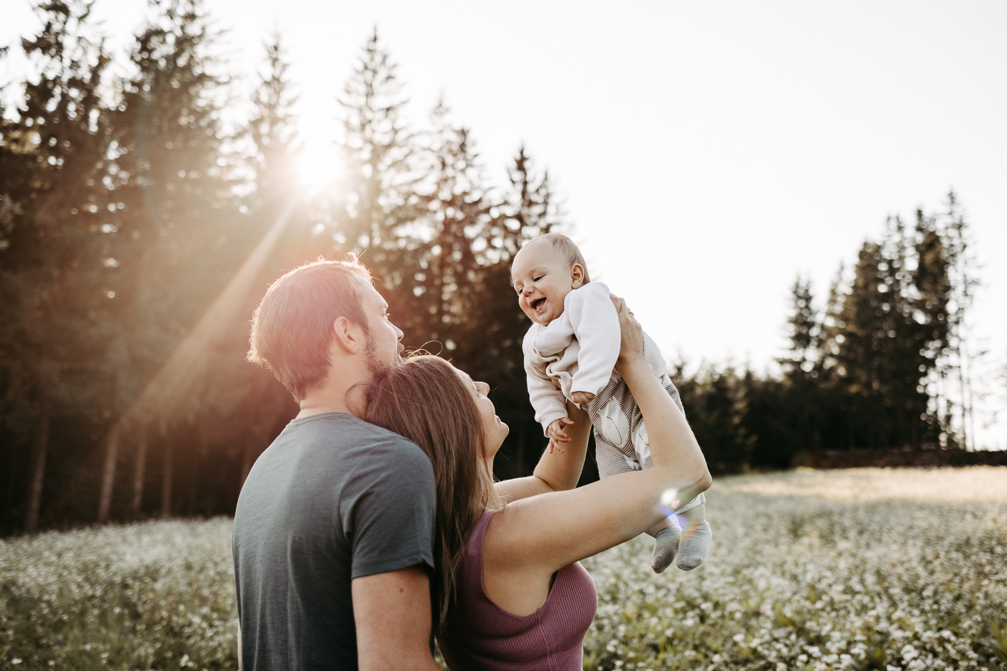 Mutter hebt lachend ihr Baby hoch, Sonnenstrahlen brechen durch die Bäume im Hintergrund