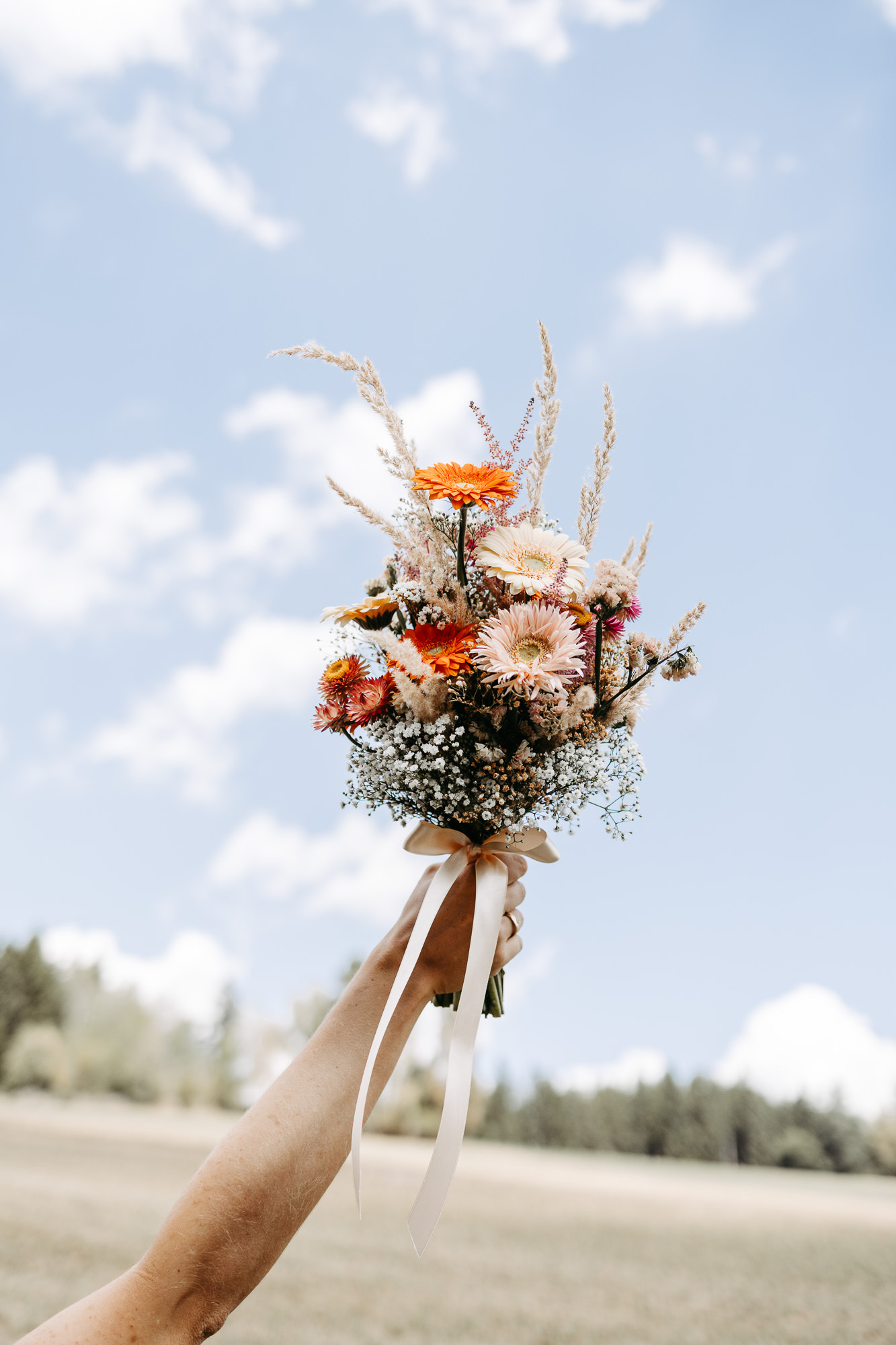 Braut hält ihren bunten Trockenblumenstrauß hoch in den blauen Himmel