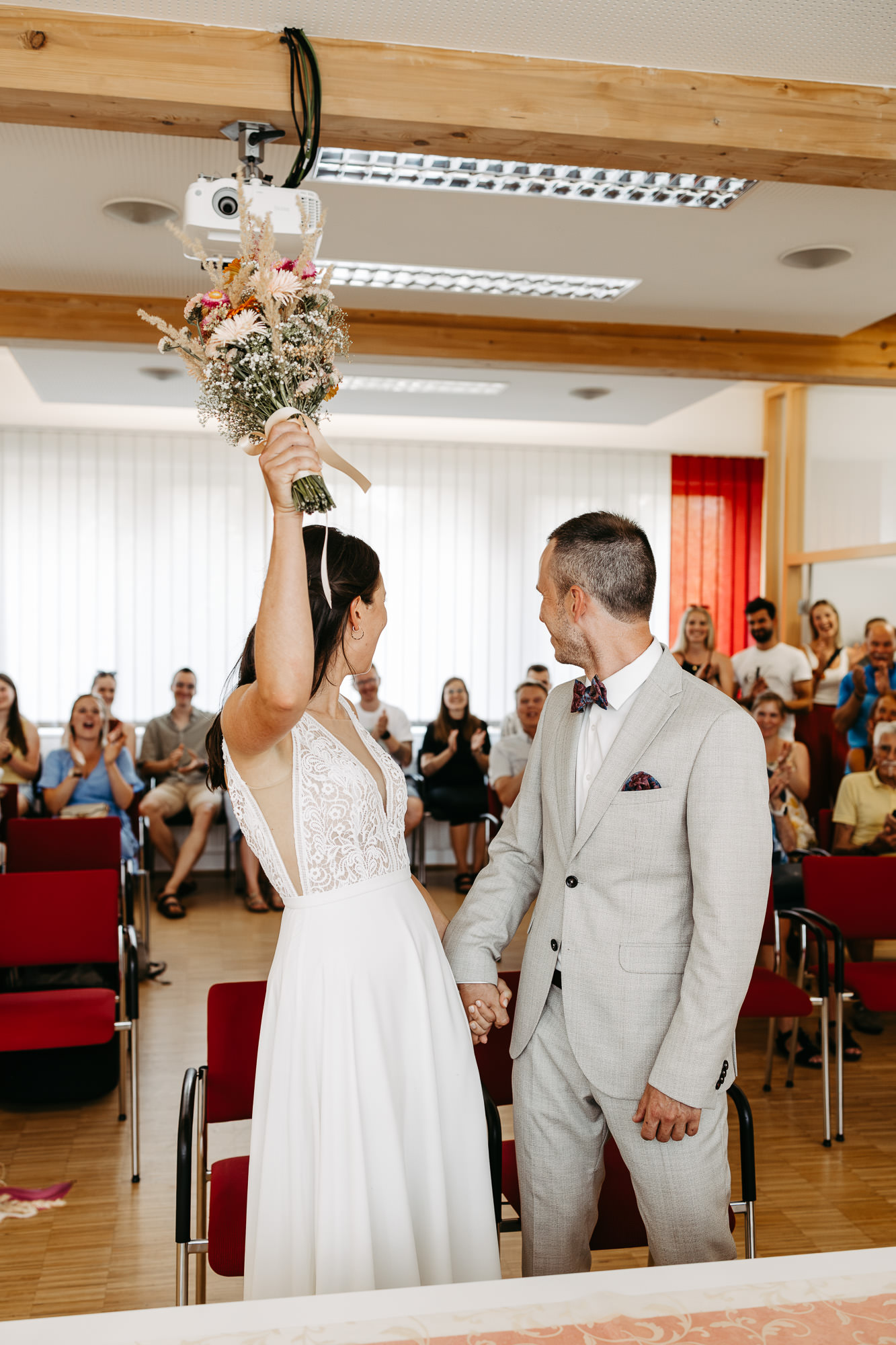 Braut jubelt mit erhobenem Blumenstrauß nach der Trauung, applaudierende Gäste im Standesamt