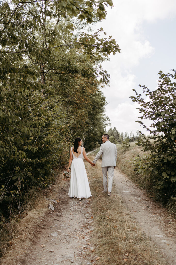 Brautpaar geht Hand in Hand einen Feldweg entlang, von hinten fotografiert