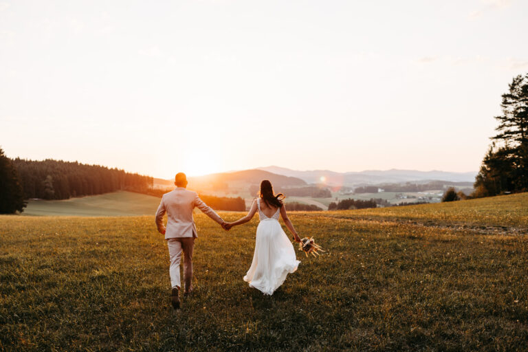 Brautpaar geht Hand in Hand über eine Wiese bei Sonnenuntergang, von hinten fotografiert