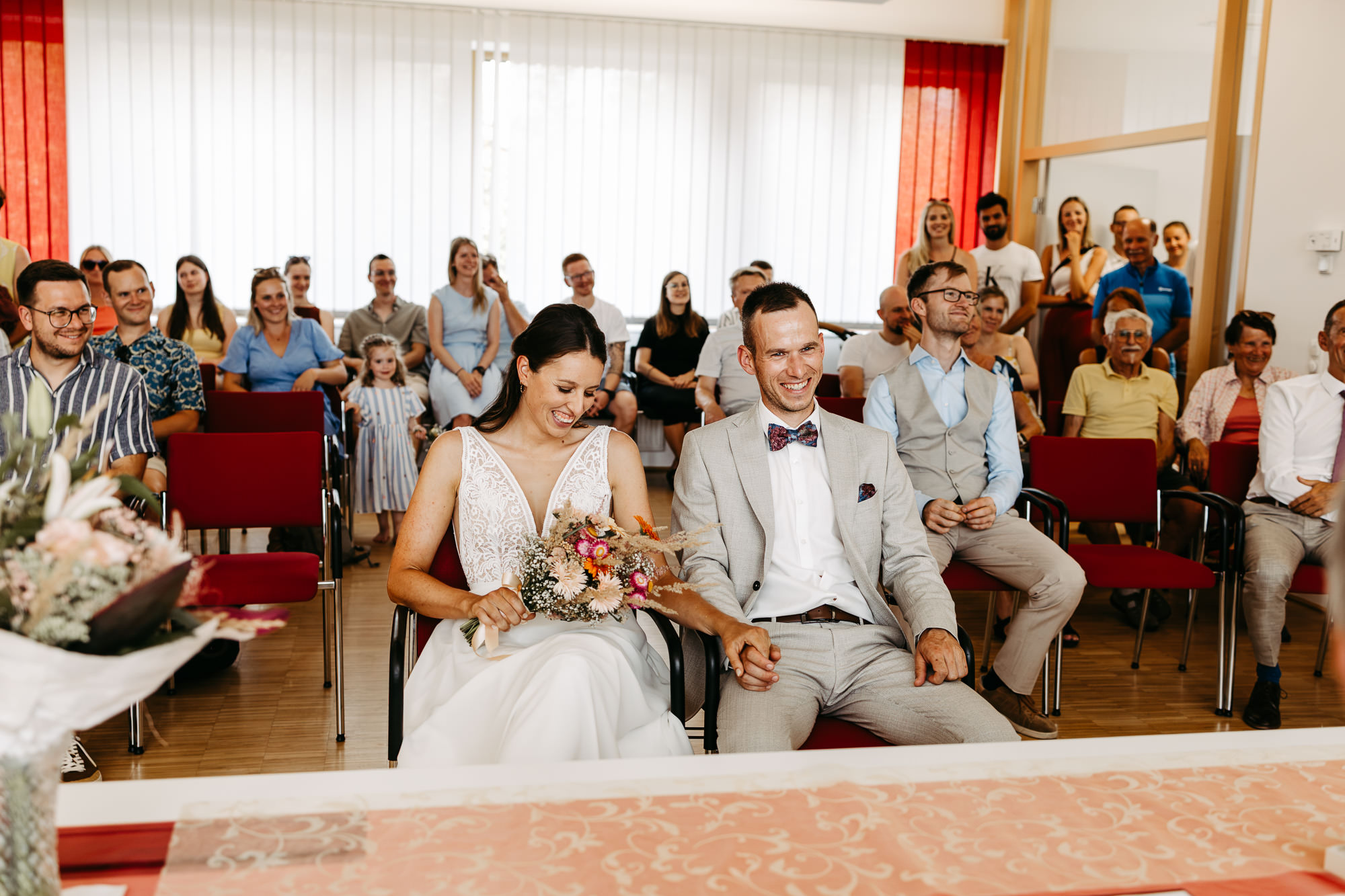 Brautpaar sitzt lachend während der Trauung im Standesamt, Gäste im Hintergrund