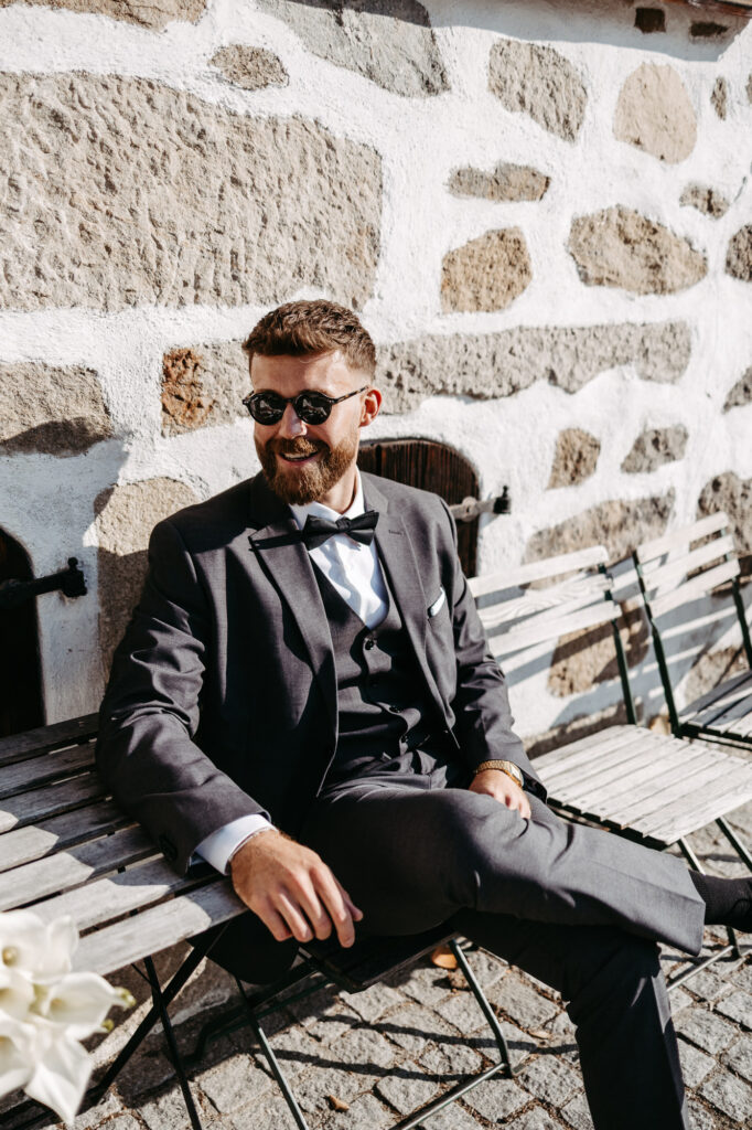 Bräutigam sitzt auf einer Bank vor einer Steinmauer, Sonnenbrille und Fliege