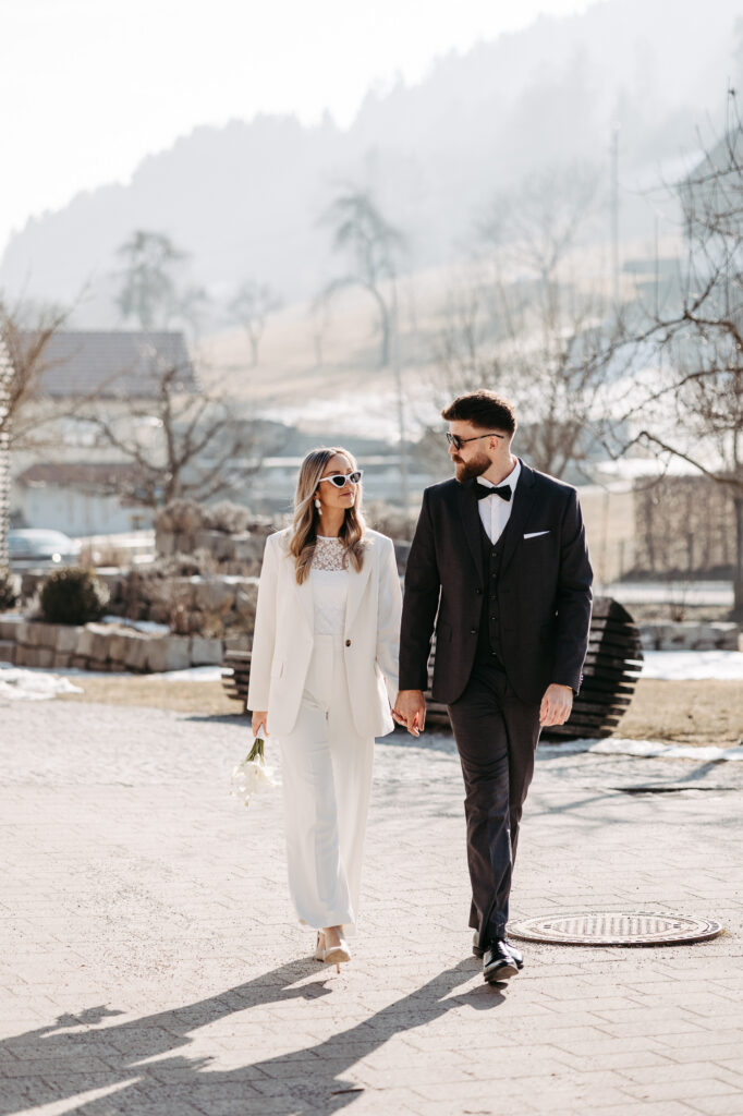 Brautpaar geht Hand in Hand durch neblige Winterlandschaft, Berge im Hintergrund, Braut im weißen Hosenanzug mit Calla-Strauß