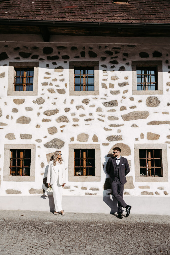 Brautpaar geht an einem alten Steinhaus mit Fensterläden entlang