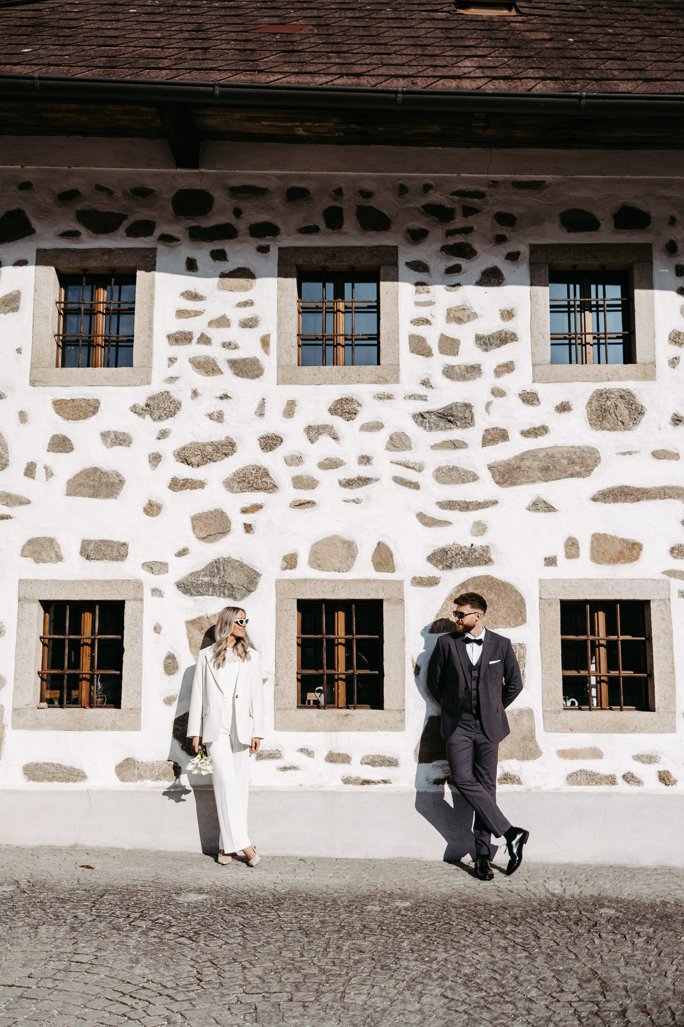 Brautpaar geht an einem alten Steinhaus mit Fensterläden entlang
