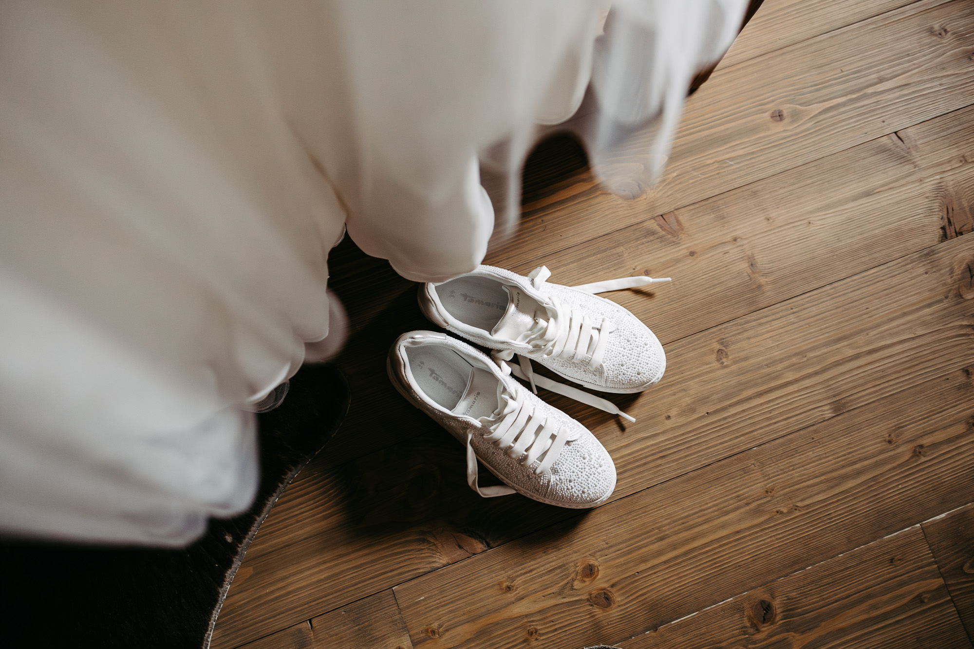 Detail: weiße Sneaker mit Strasssteinen auf Holzboden, Brautkleid im Hintergrund