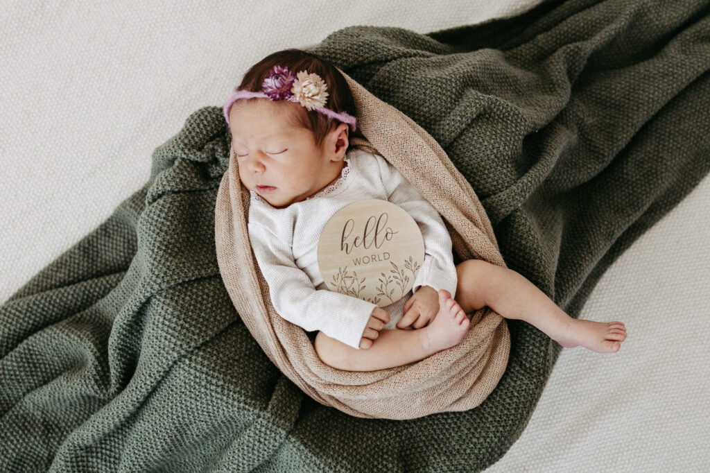 Baby eingewickelt in eine Decke mit „Hello World"-Holzschild und rosa Blumen-Haarband