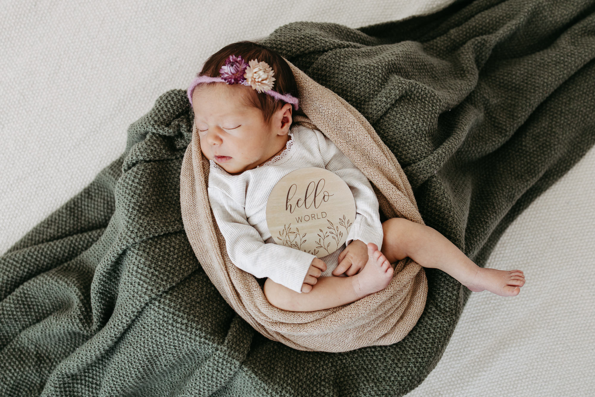 Baby eingewickelt in eine Decke mit „Hello World"-Holzschild und rosa Blumen-Haarband