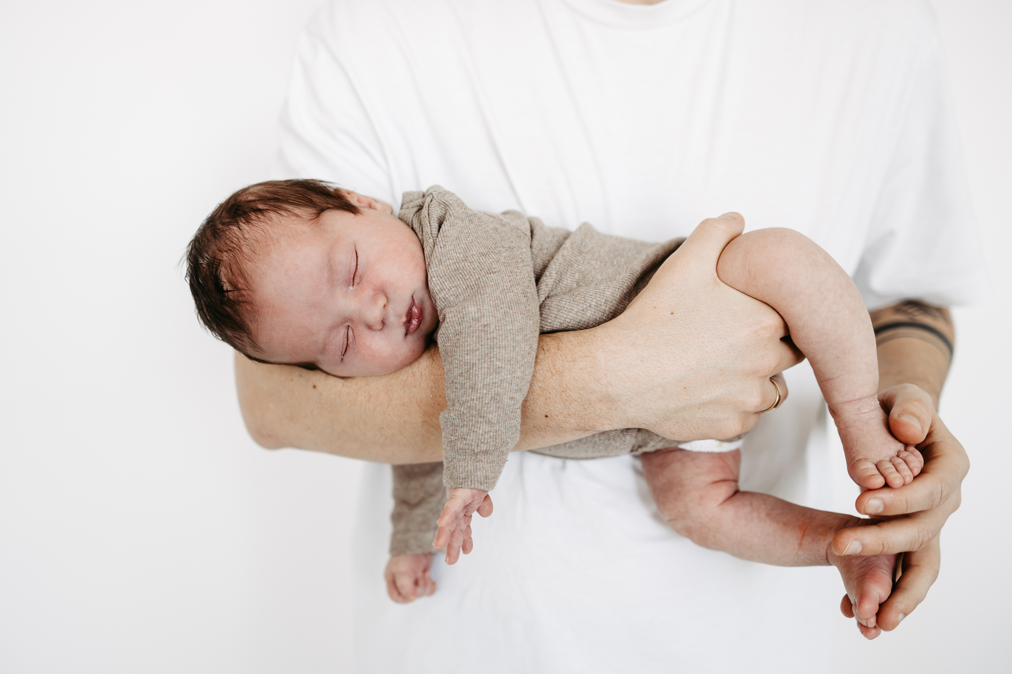 Baby schläft in beigem Strickoutfit auf Vaters Arm gebettet, weißer Hintergrund