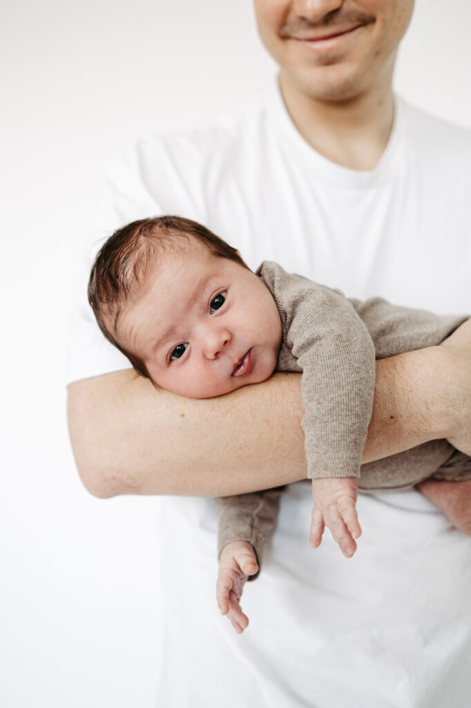 Baby liegt auf Vaters Arm, blickt zur Kamera, weißer Hintergrund