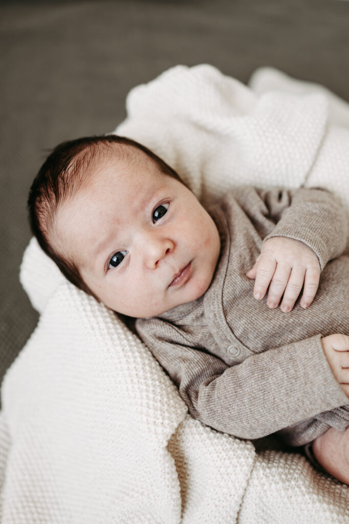 Baby liegt wach mit großen Augen auf cremefarbener Wolldecke, graues Strickoutfit