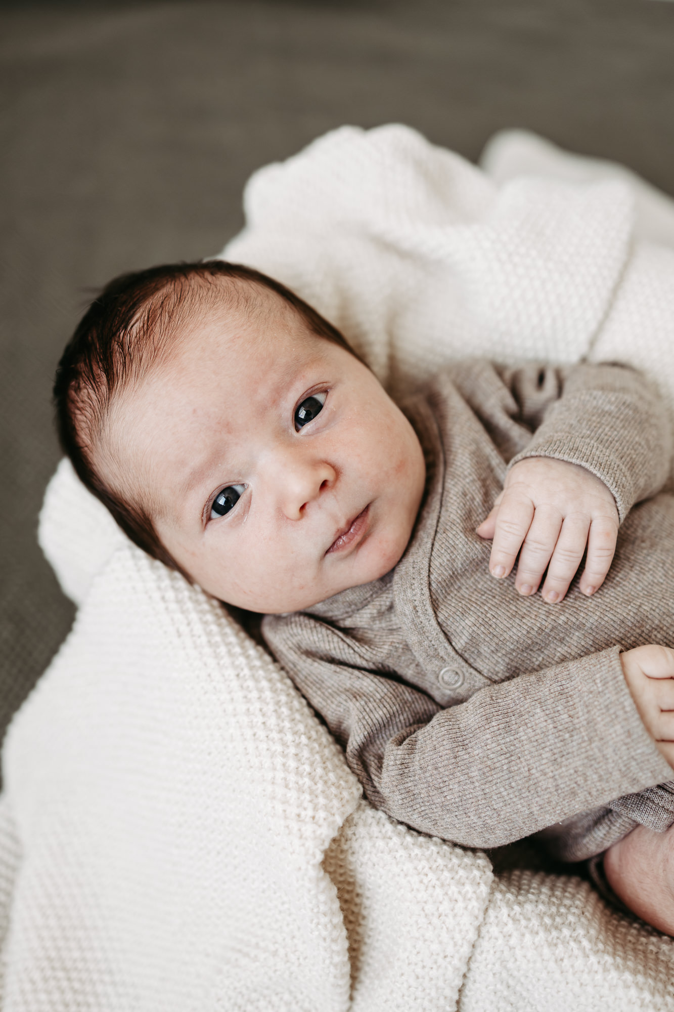 Baby liegt wach mit großen Augen auf cremefarbener Wolldecke, graues Strickoutfit