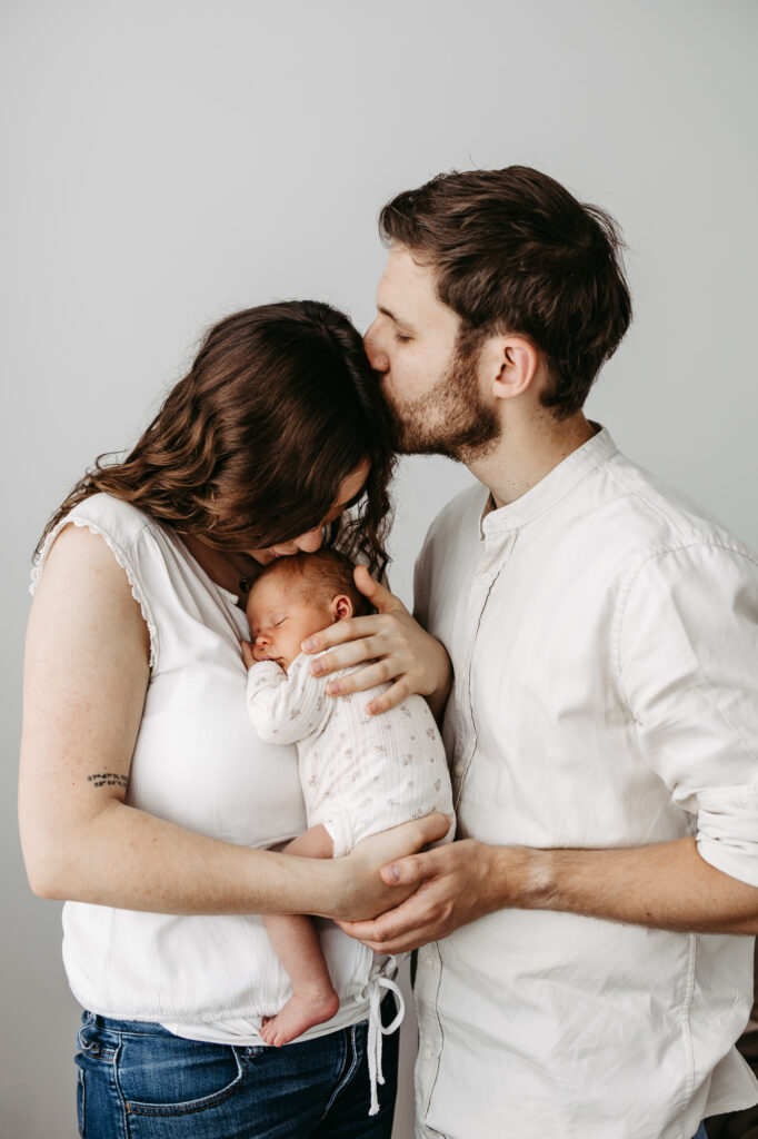 Eltern am Fenster, der Vater küsst die Stirn der Mutter, während sie das schlafende Baby hält