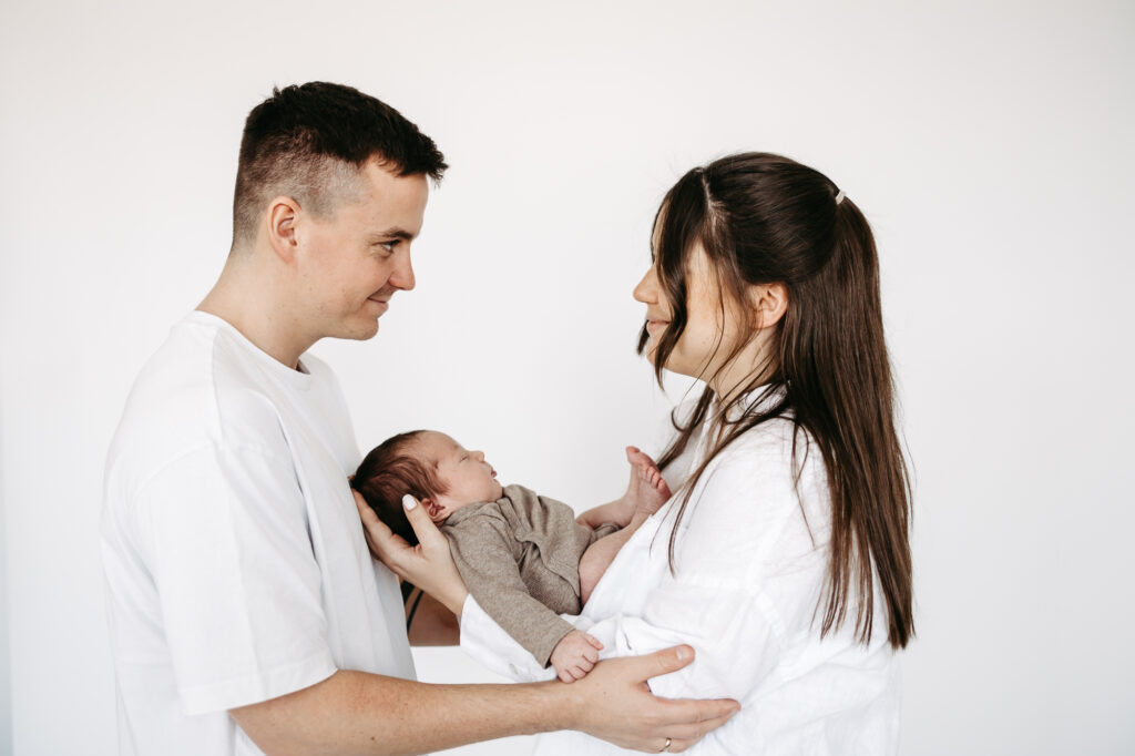 Eltern stehen sich im Profil lächelnd gegenüber, Baby gehalten zwischen ihnen