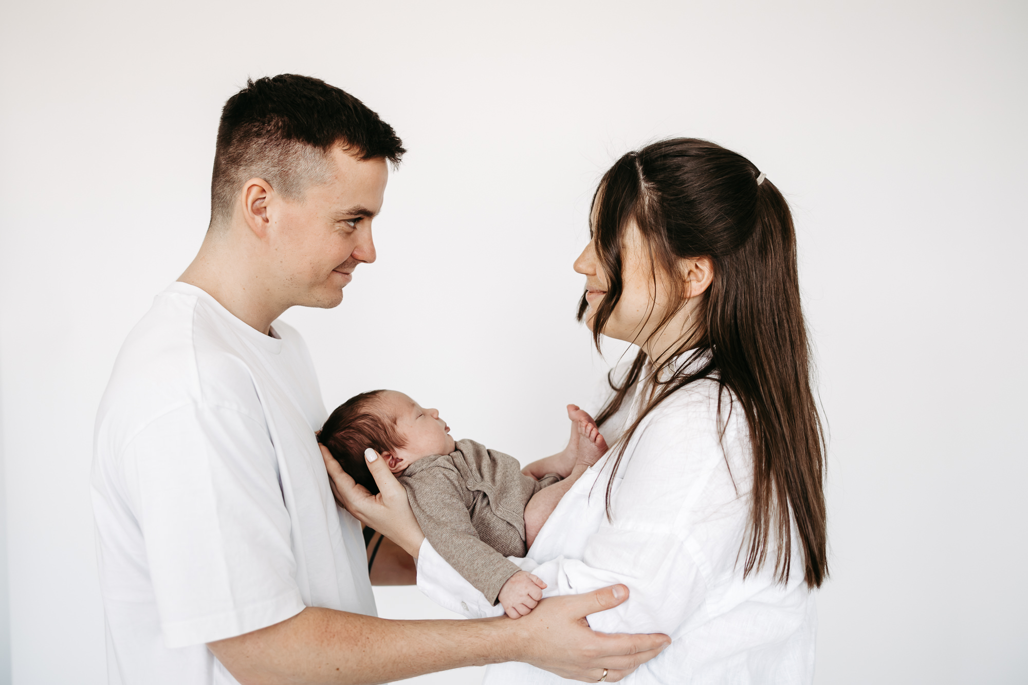 Eltern stehen sich im Profil lächelnd gegenüber, Baby gehalten zwischen ihnen