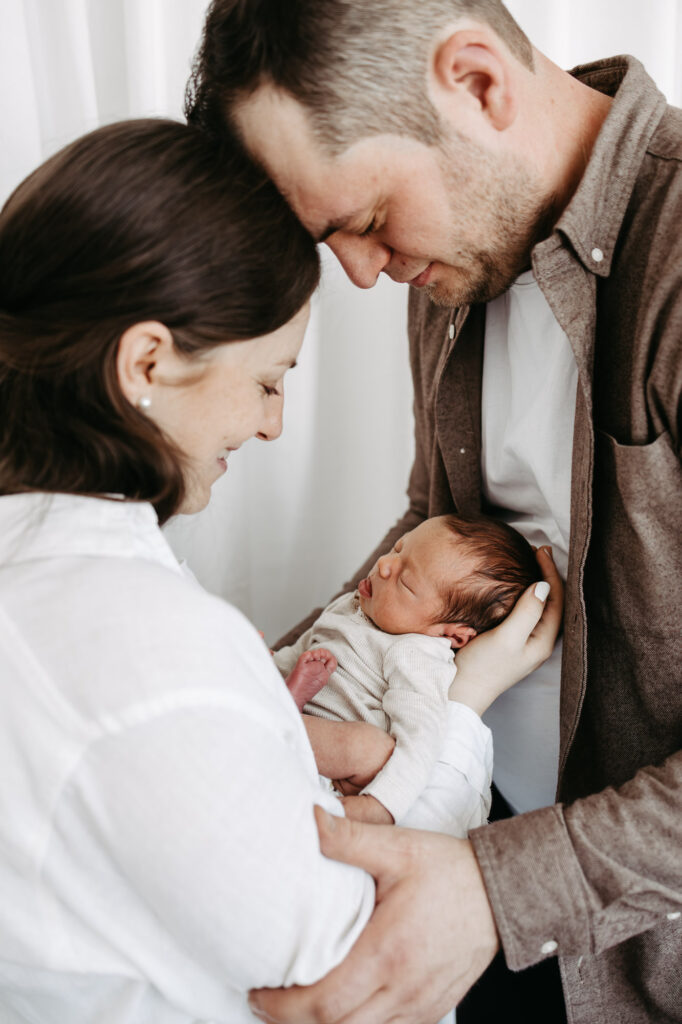 Eltern berühren sich Stirn an Stirn, das Baby liegt dazwischen im Arm