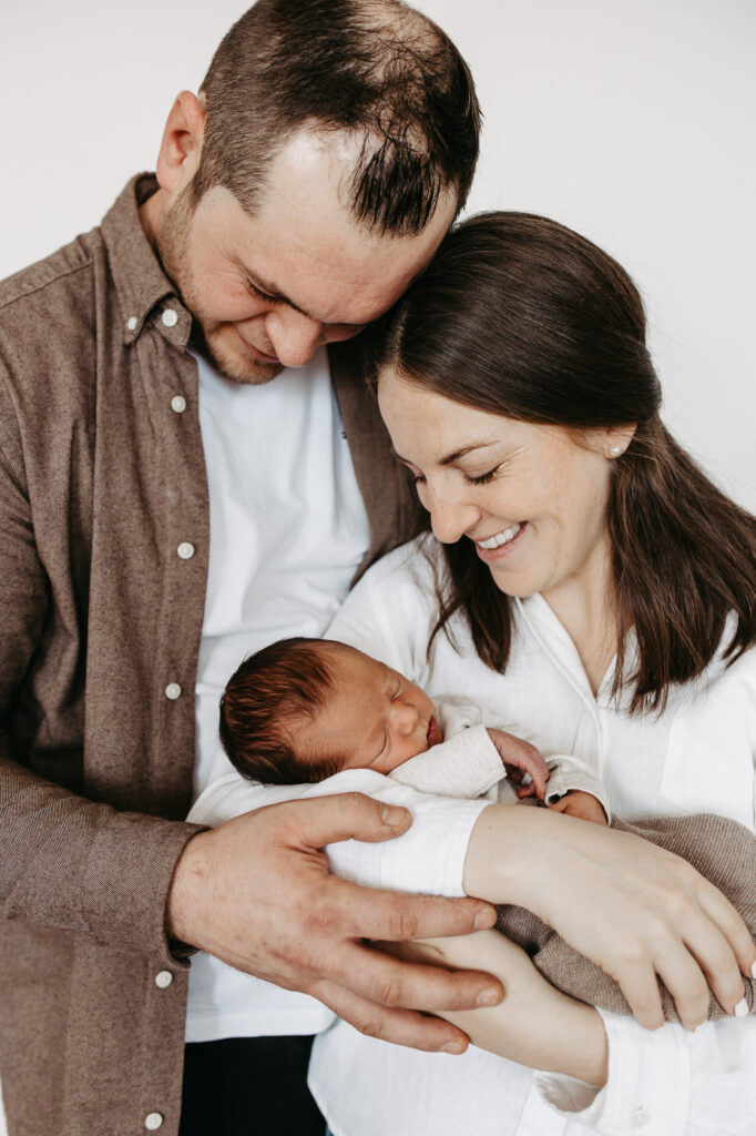 Eltern berühren sich Stirn an Stirn und lächeln, der Vater trägt das eingewickelte Baby