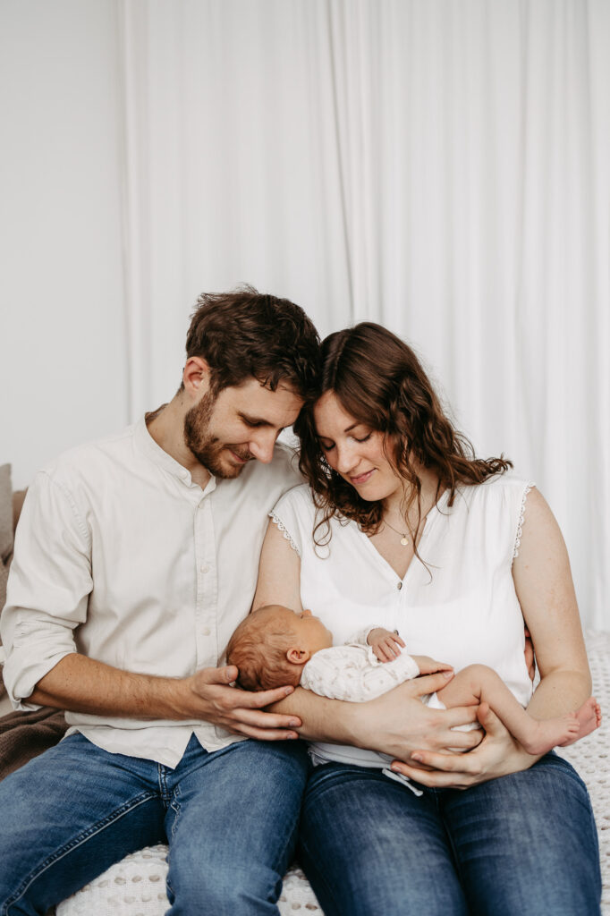 Eltern stehen am Fenster, lächelnd blicken sie auf ihr Baby im Arm der Mutter