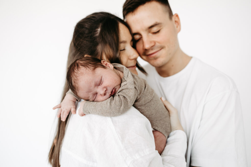 Eltern lehnen Wange an Wange, Baby schläft zwischen ihnen