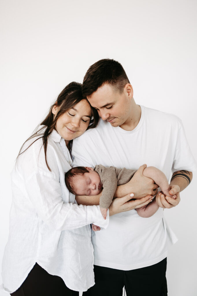 Familie steht eng zusammen, Vater küsst Mutter auf die Stirn, Baby schläft dazwischen