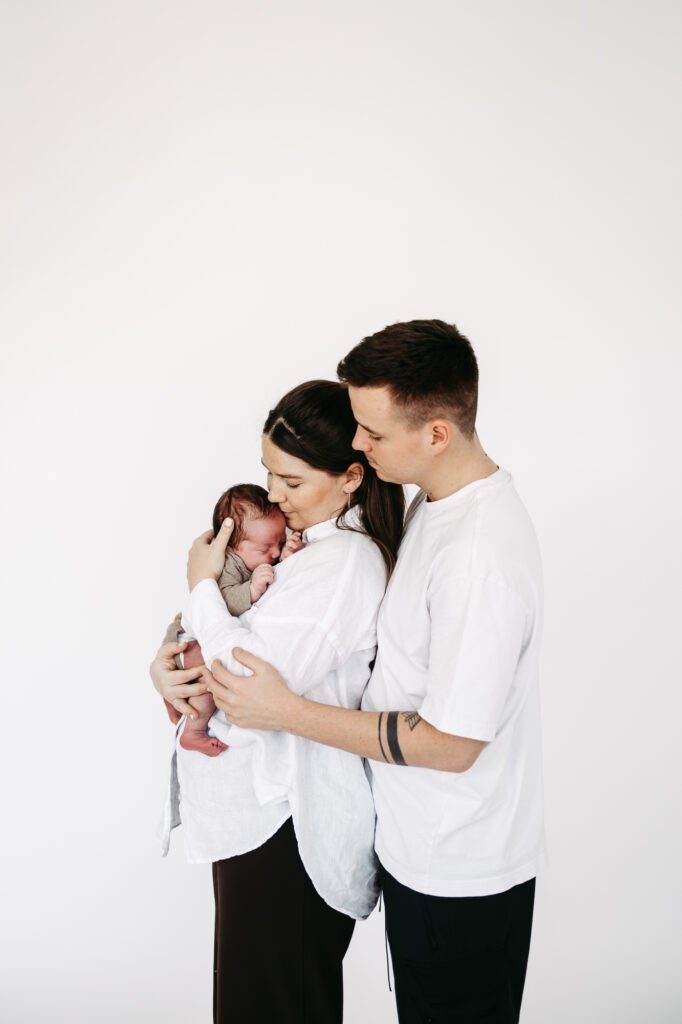 Familienportrait: Mutter hält das eingewickelte Baby, Vater umarmt beide von hinten, weißer Hintergrund