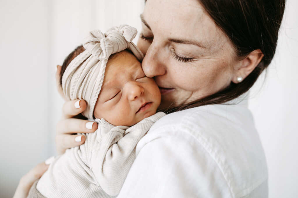 Mutter küsst die Stirn ihres Babys, das ein cremefarbenes Haarband mit Schleife trägt
