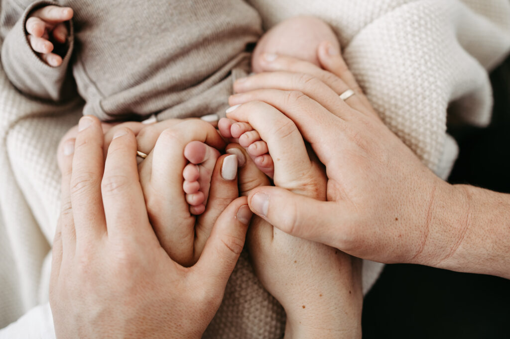 Nahaufnahme: Hände der Eltern halten die Füßchen des Babys, Eheringe sichtbar