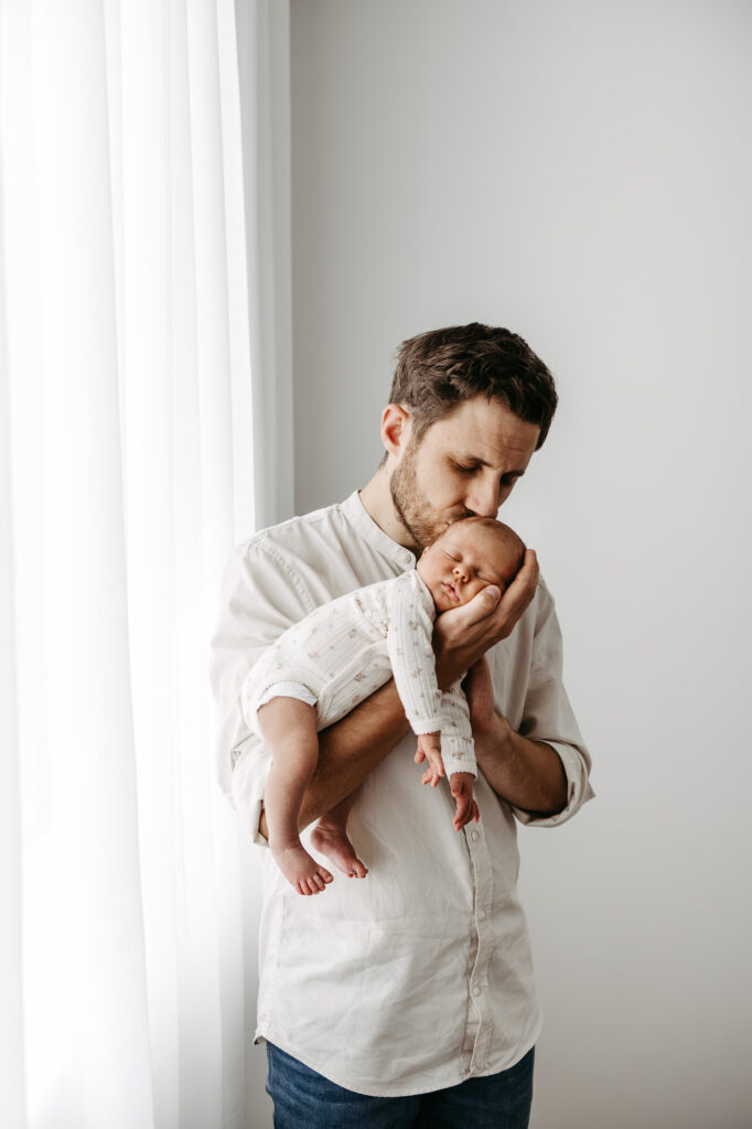 Vater steht am Fenster und küsst den Kopf seines Babys